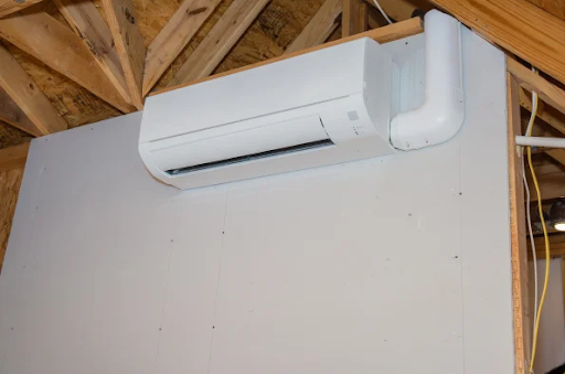 A white wall-mounted ductless mini-split air handler is positioned on a grey drywall sheet under exposed wooden roof trusses, featuring an attached white line set cover.