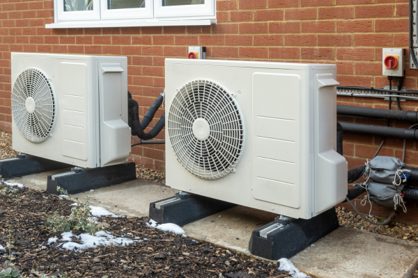 Two white outdoor heat pump condenser units are installed on concrete mounting pads along a red brick wall with visible insulated refrigerant lines and electrical connections.