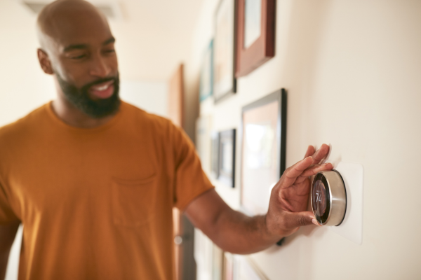 A man with a beard and an orange shirt smiles while turning the dial of a circular smart thermostat mounted on a wall decorated with framed pictures.