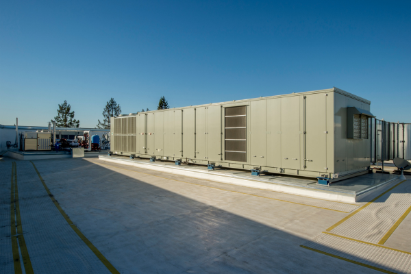A massive industrial-grade air handling unit is installed on a commercial flat roof with support rails and visible external components under a clear sky.