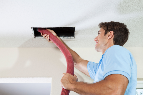 A service technician inserts a large red vacuum hose into a rectangular ceiling vent to remove debris from a residential air duct system.