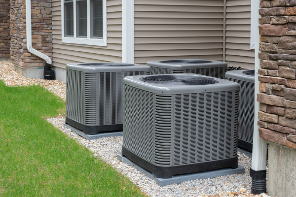 Four grey outdoor air conditioning or heat pump condenser units are installed in a row on concrete pads along the side of a residential building.