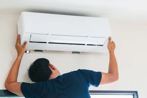 A technician in a dark blue shirt carefully mounts a white ductless mini-split air handler unit onto a wall bracket inside a home.