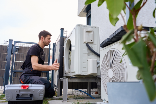 A technician in dark workwear kneels next to an outdoor heat pump unit with an open service panel and a grey tool box nearby.