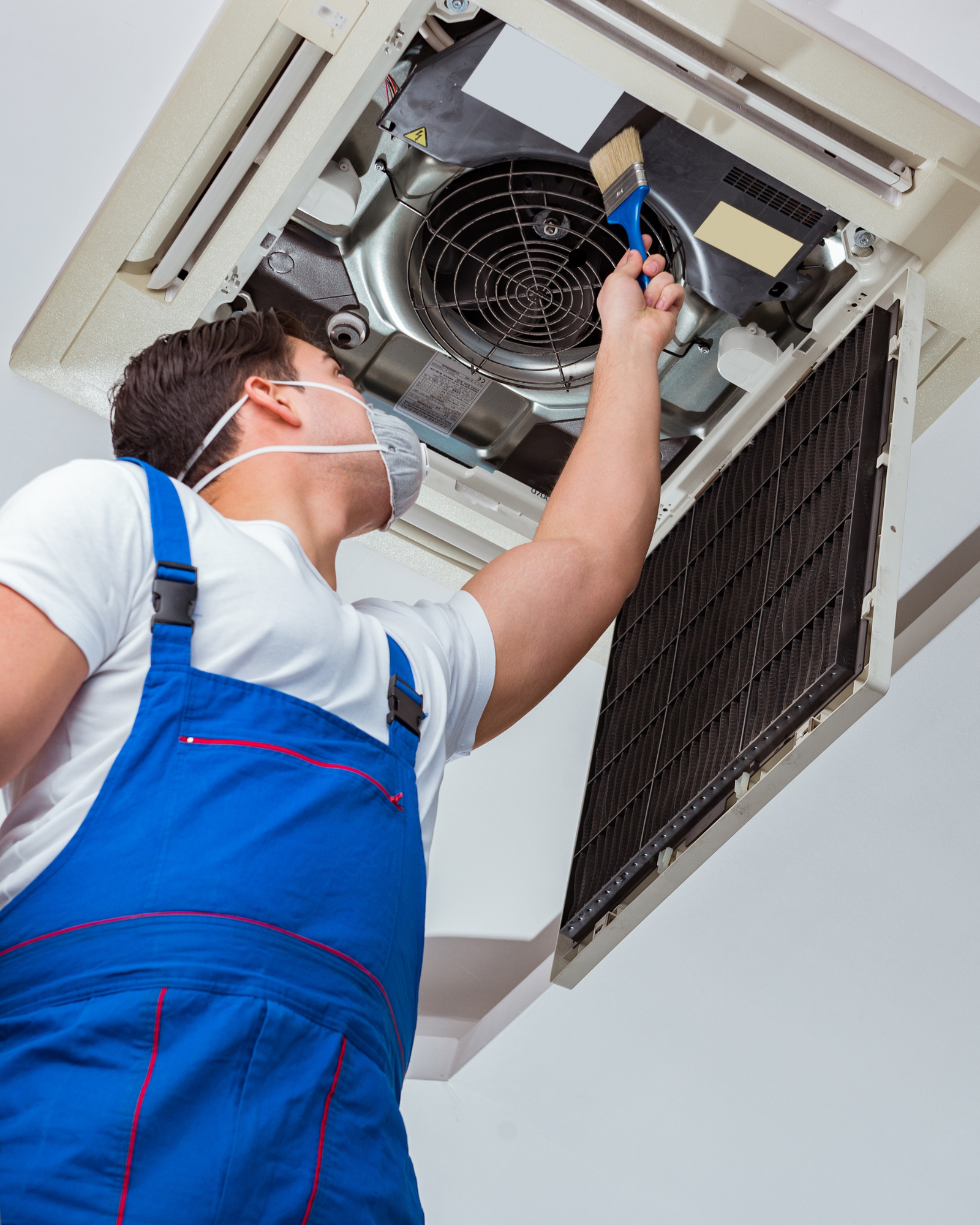 A technician wearing a protective face mask and blue overalls uses a small brush to clean the internal fan and components of an open ceiling-mounted cassette air conditioning unit.