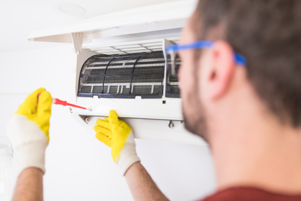 A technician wearing a blue uniform and a face mask carefully fits the decorative front panel onto a ceiling-mounted cassette air handler during a professional installation.