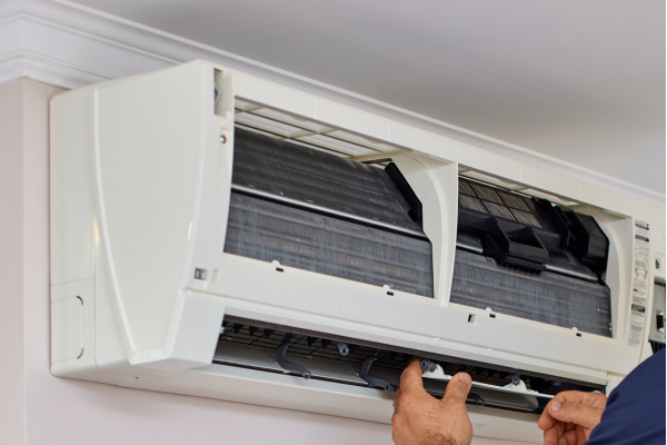 A close-up view of a technician's hands carefully removing or adjusting the lower access panel of an indoor wall-mounted air handling unit to inspect the internal cooling coils.