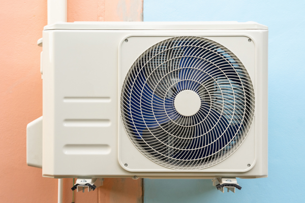 A wall-mounted white outdoor condenser unit with a large circular fan grille is installed against a dual-colored peach and blue exterior wall.