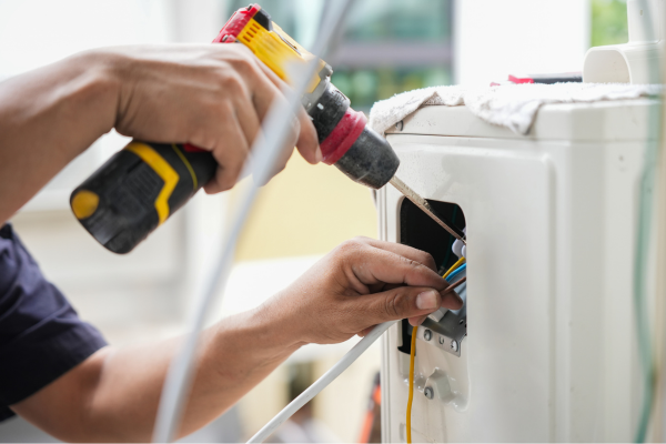 A close-up view of a technician's hands using a yellow power drill to secure electrical connections and colored wires within the access panel of an outdoor air conditioning unit.