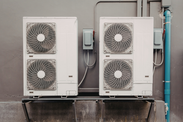Two large, white outdoor HVAC condenser units are mounted side-by-side on a metal rack against a grey wall, each featuring individual electrical safety disconnect boxes and conduit wiring.