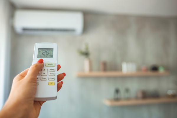 A person’s hand holds a white digital remote control in the foreground to adjust the settings of a wall-mounted ductless air conditioner visible in the blurred background.