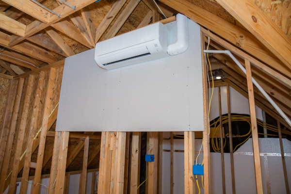 A white wall-mounted ductless air handling unit with connected white refrigerant lines is installed on a drywall panel within an unfinished attic space showing wooden rafters and electrical wiring.