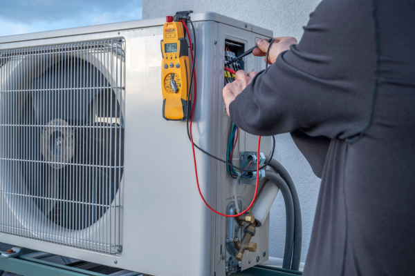 A technician uses a digital multimeter to measure electrical current and troubleshoot the wiring inside the service panel of an outdoor air conditioning condenser.