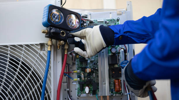 A HVAC technician wearing protective gloves uses manifold gauges to inspect and service an outdoor air conditioning unit with the access panel open, showing internal electrical components during maintenance.