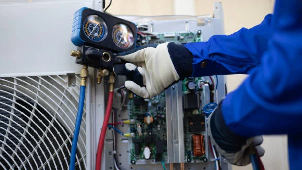 A HVAC technician wearing protective gloves uses manifold gauges to inspect and service an outdoor air conditioning unit with the access panel open, showing internal electrical components during maintenance.