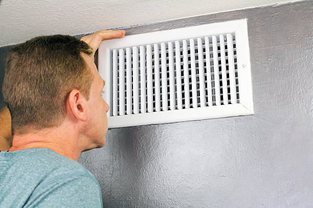 A homeowner inspecting a residential HVAC air vent mounted on an interior wall to check airflow and indoor ventilation performance.