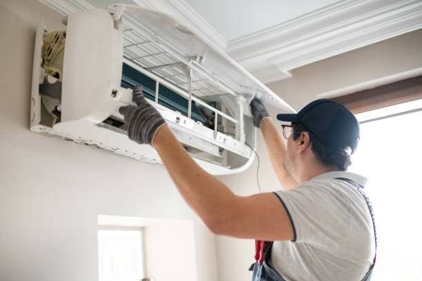A technician in a dark cap, glasses, and gray gloves reaching up to open the front panel of a wall-mounted indoor air conditioning unit for inspection or maintenance.