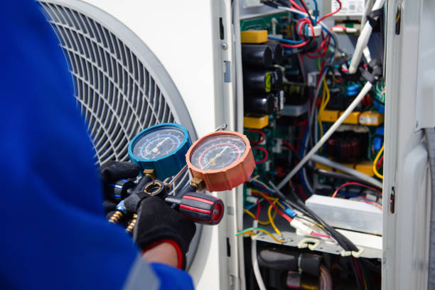 A technician in a blue uniform and black gloves holding blue and red refrigerant pressure gauge manifolds in front of an open outdoor AC unit with visible internal wiring and components.