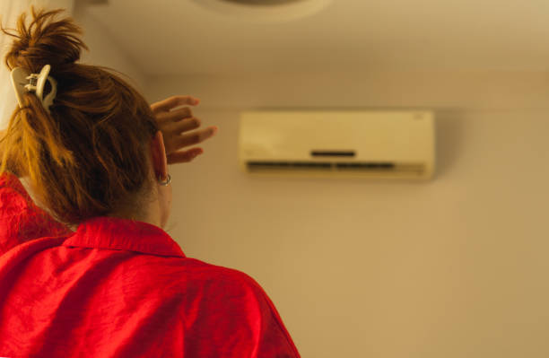 A woman in a red robe with her hair up fanning herself with her hand while standing in front of a wall-mounted AC unit that appears to not be cooling the room effectively.