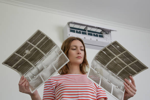A woman in a red striped shirt holding two heavily dust-clogged air conditioning filters with both hands, standing beneath an open wall-mounted AC unit indoors.