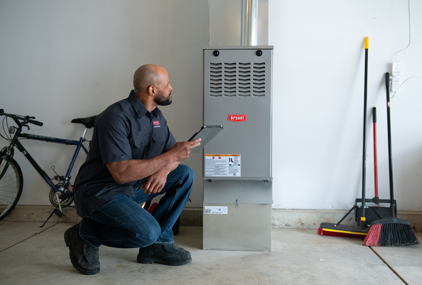 An ATX Heating & Air Conditioning technician crouching next to a gray Bryant gas furnace in a garage while holding a tablet for diagnostics.