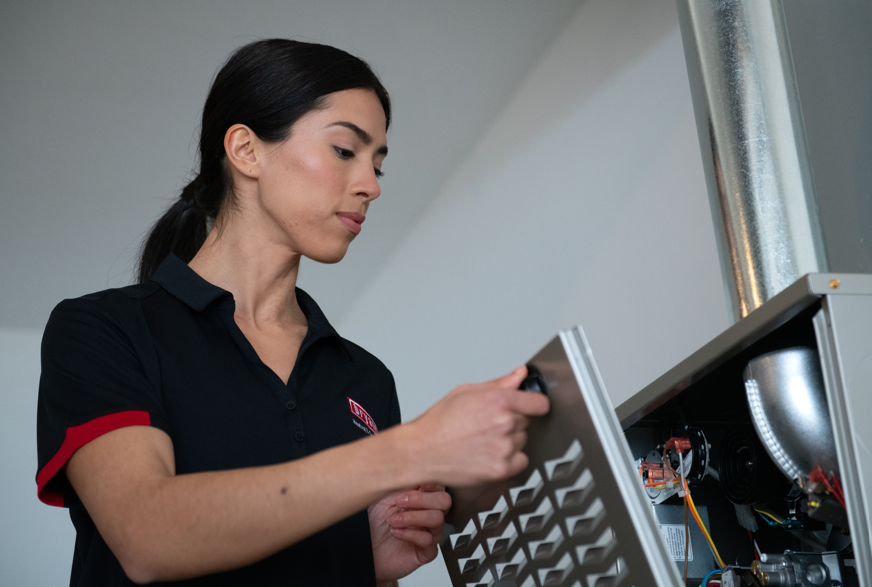 A female ATX Heating & Air Conditioning technician removing the louvered front panel of a Bryant gas furnace to inspect the internal wiring and components.