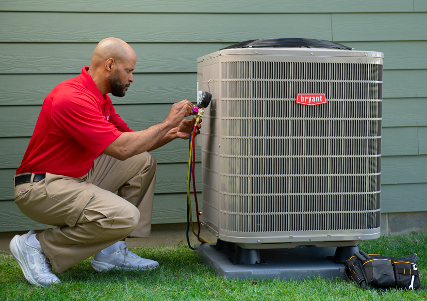 An ATX Heating & Air Conditioning technician in a red shirt using manifold gauges to test an outdoor Bryant heat pump unit.