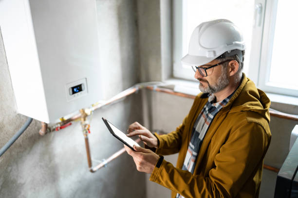 Inspector wearing a hard hat and glasses reviewing a tablet while standing beside a wall-mounted boiler with exposed copper piping.
