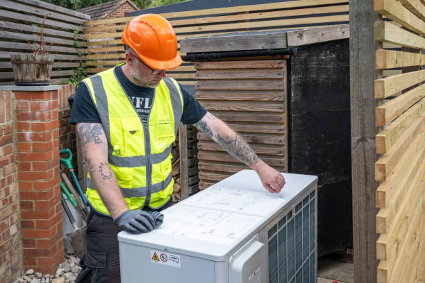 Technician wearing an orange hard hat and safety vest inspecting the top of an outdoor air conditioning unit in a backyard.