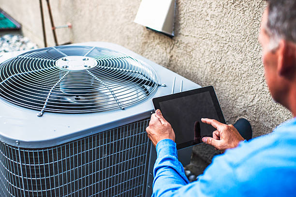 Technician using a tablet to record inspection notes beside an outdoor air conditioning condenser unit.