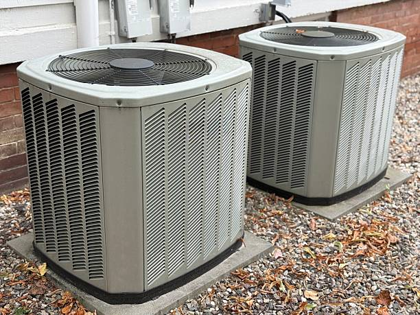 Two outdoor air conditioning condenser units installed side by side on a gravel bed against a house exterior."
