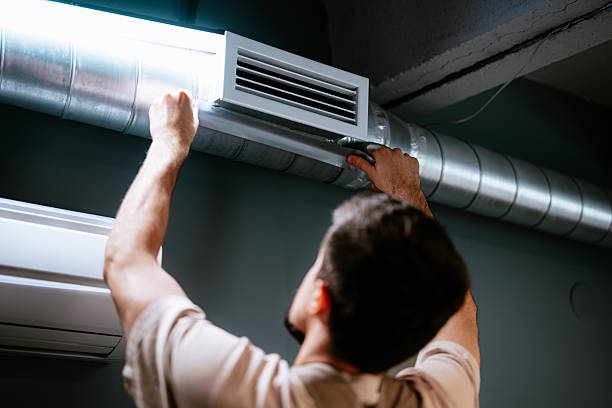 HVAC technician installing a vent grille on exposed round metal ductwork next to a wall-mounted AC unit